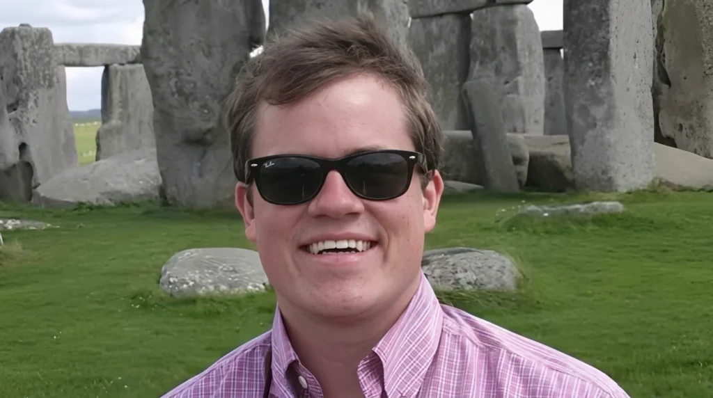 Jett Puckett wearing sunglasses and a pink button-up shirt smiling with Stonehenge in the background